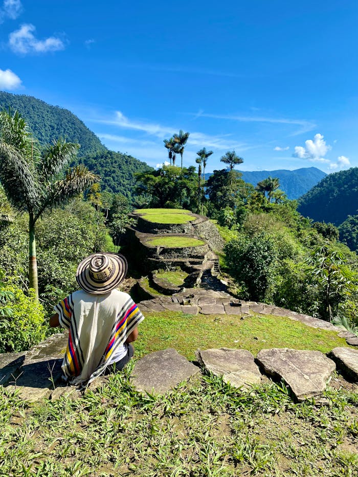 A person in traditional attire sits overlooking the ancient terraces of Colombias Lost City in lush jungle surroundings.